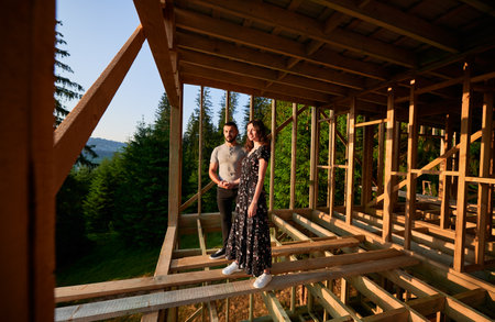 Man and woman inspecting their future wooden frame house nestled in the mountains near forest. Youthful couple at construction site in early morning. Concept of contemporary ecological construction.の写真素材