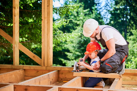 Father with toddler son building wooden frame house near forest. Male builder teaching his son how to cut board using electric saw on construction site, wearing helmets and overalls on sunny day.の写真素材