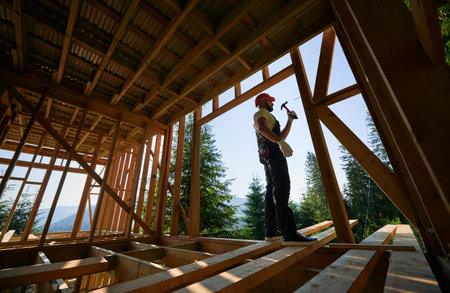 Carpenter constructing wooden frame, two-story house near forest. Bearded man hammering nails with hammer while dressed in protective helmet. Concept of environmentally-friendly modern construction.の写真素材