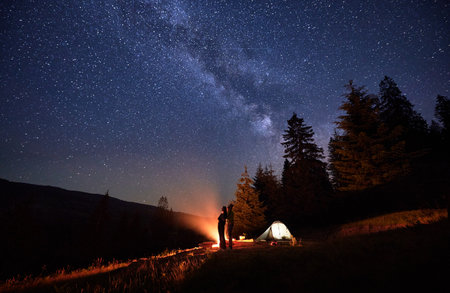Overnight camping expedition, under shimmering stars. Man and woman intrepid hikers near bonfire and tent pitched near woods, under resplendent night sky with Chumack Path as their celestial guide.の写真素材