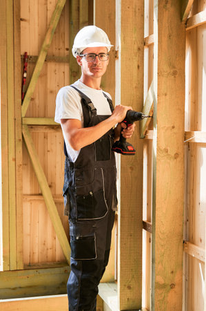 Carpenter constructing wooden framed house. Man worker in glasses working with screwdriver, wearing work overalls and helmet. Concept of modern eco-friendly construction.の写真素材