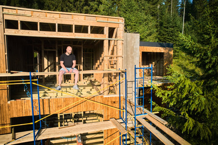 Designer is working on wooden frame two-story house construction site near the forest, sitting on scaffold. The concept behind it is modern and environmentally friendly construction.の写真素材