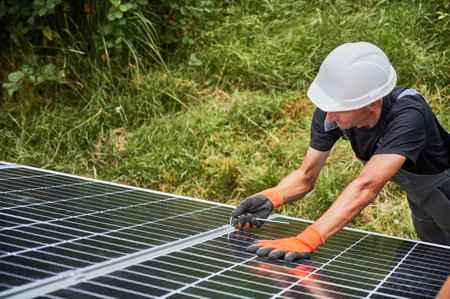 Male worker mounting photovoltaic solar panel system outdoors. Man engineer placing solar module on metal rails, wearing construction helmets and work gloves. Renewable and ecological energy.の写真素材