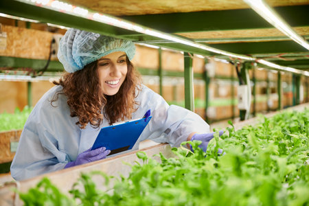 Joyful woman checking plant growth and taking notes in greenhouse. Female researcher in disposable cap and garden gloves looking at green leafy plants and smiling while writing on clipboard.の写真素材