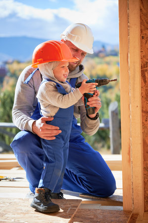 Father with toddler son building wooden frame house. Happy boy helping his daddy, using screwdriver on construction site, wearing helmet and blue overalls on sunny day. Carpentry and family concept.の写真素材