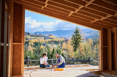 Father, mother and son building wooden frame house. Toddler boy helping his daddy, while woman looking for them on construction site. Guys wearing blue overalls. Carpentry, family concept.の写真素材