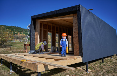 Boy toddler playing as builder on construction site. Child carpenter learning to build wooden frame house in the Scandinavian style barnhouse outdoor on sunny day. Carpentry and workshop concept.の写真素材