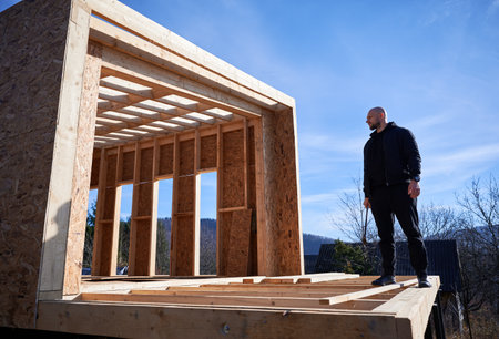Male carpenter building wooden frame house in the Scandinavian style barnhouse. Bald man standing on construction site, inspecting quality of work on sunny day with blue sky on background.の写真素材