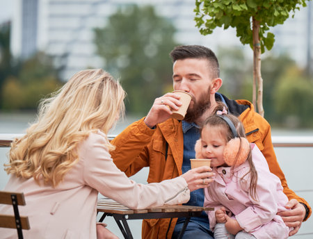 Young family man and woman drinking of coffee while spending time with daughter in outdoor cafe. Happy husband and wife sitting at the table with drink cups in new urban district.の写真素材