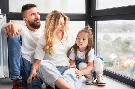 Family portrait of man, woman and little girl in newly built apartment. Bearded man looking out the window while sitting on the floor next to wife and daughter. Family and new home concept.の写真素材