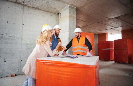 Family with child discussing architectural plan of new apartment with construction worker, standing inside apartment building under construction and talking with builder.の写真素材