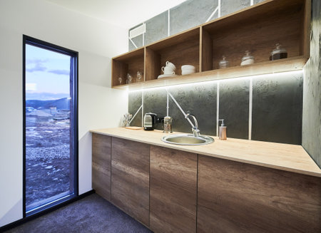 Modern kitchen with white walls and glass door with scenic view inside. Side view of kitchen area with wooden shelves and working surface, illuminated with warm LED light indoor. Concept of interior.の写真素材