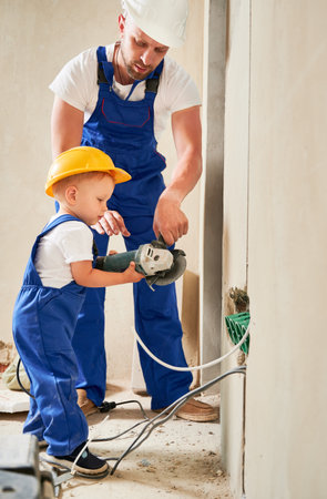 Child holding electric angle grinder while installing cable canals and sockets in wall with father. Man and kid in safety construction helmet using power tool and mounting electrical wiring at home.の写真素材