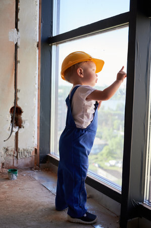 Full length of adorable little boy looking out the window in apartment under renovation. Kid construction worker wearing safety helmet and work overalls. Concept of childhood and home repair.の写真素材