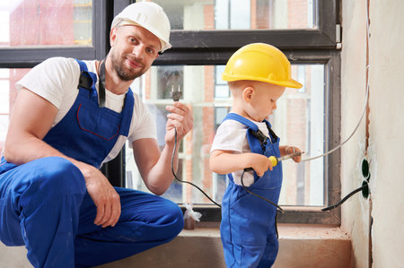 Male electrician holding electrical plug and smiling while child using electric soldering iron tool. Man and kid wearing work overalls and safety helmets while working on home renovation.の写真素材