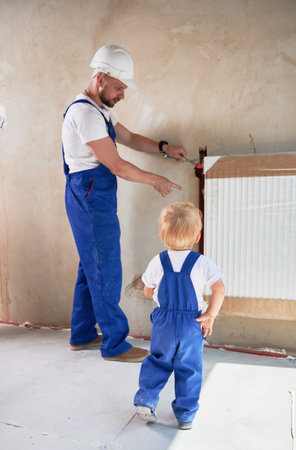 Male plumber using wrench tool and pointing at radiator while installing heating system together with child. Man and baby boy wearing work overalls while working on home renovation.の写真素材