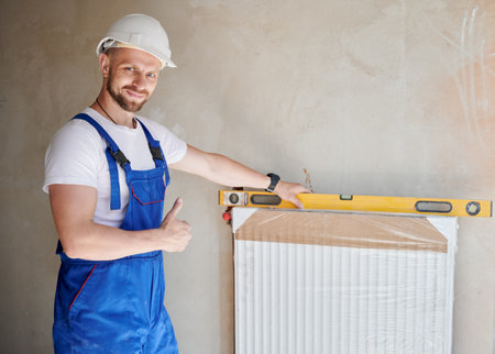 Male worker installing heating radiator in apartment under renovation, looking at camera and showing thumbs up gesture. Man in work overalls and helmet using spirit level ruler tool.の写真素材