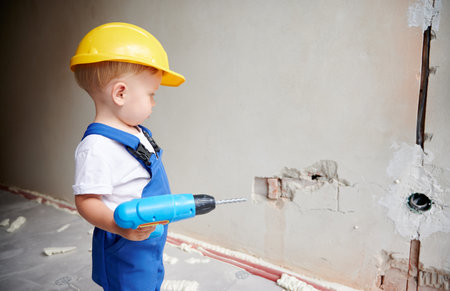 Baby boy construction worker with toy electric drill looking at the wall in apartment. Kid in safety helmet and work overalls holding cordless power tool for repair works. Home renovation concept.の写真素材