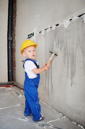 Full length of baby boy holding hammer and looking at camera while standing by the wall. Kid wearing safety helmet and work overalls while using construction tool at home during renovation.の写真素材