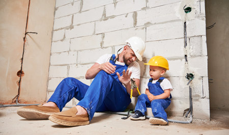Male builder in work overalls showing pliers to little boy in apartment under renovation. Man and kid in safety helmets sitting by brick wall in empty building. Home renovation and parenting concept.の写真素材