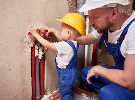 Male plumber and child measuring pipes level with building instrument. Man and little boy in safety construction helmet using spirit level tool while installing plumbing pipes in apartment.の写真素材
