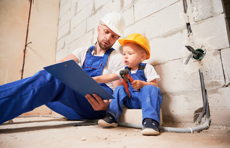 Male worker sitting by brick wall and pointing at clipboard while little boy holding tool. Man and kid wearing safety helmets and work overalls while working with documents at construction site.の写真素材