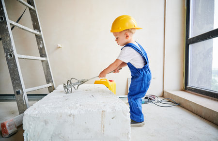 Full length of little boy construction worker using building instrument while working on home renovation. Kid in work overalls and safety helmet preparing material for repair and renovation works.の写真素材