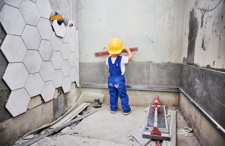 Little boy checking wall surface with spirit level in apartment. Back view of kid in work overalls and safety construction helmet using level instrument while working on home renovation.の写真素材