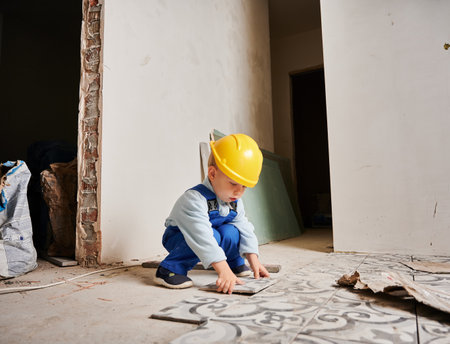 Little boy construction worker installing ceramic tile on the floor in apartment under renovation. Kid wearing safety helmet and work overalls while playing at home.の写真素材