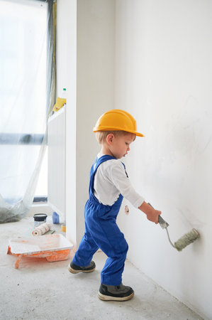 Little boy painting wall in apartment under renovation. Kid in safety helmet and work overalls using paint roller while playing at home under renovation.の写真素材