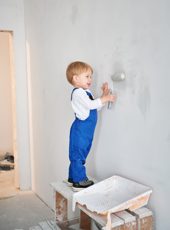 Full length of smiling little boy construction worker painting wall in apartment under renovation. Adorable kid in work overalls using paint roller while working on home renovation.の写真素材