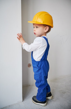 Child construction worker standing near the wall in apartment under renovation. Kid in safety helmet and work overalls playing at future home.の写真素材