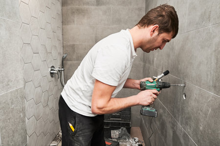 Male worker using electric drilling machine and construction brush while working on bathroom renovation. Male plumber drilling wall with power drill while installing shower system at home.の写真素材