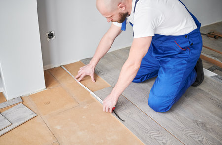 Male construction worker using tape measure while installing laminate flooring in apartment under renovation. Man in workwear measuring distance from wall to laminate board with measuring tape ruler.の写真素材