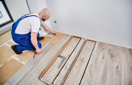 Male builder checking distance from wall to laminate board. Man in work overalls using tape measure while installing laminate flooring in apartment under renovation. Hardwood floor renovation concept.の写真素材