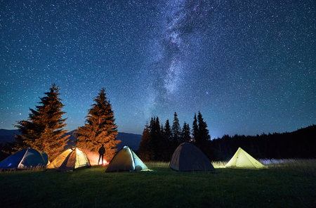 Night camping in mountains under starry sky. Silhouette of person standing in campsite near tents, burning campfire under beautiful sky with stars and Milky way. Concept of tourism, beauty of nature.の写真素材