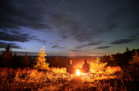 Happy couple staring at starry sky. Young people sitting in chairs and warming by campfire. Travelling couple having a rest together. Night camping.の写真素材