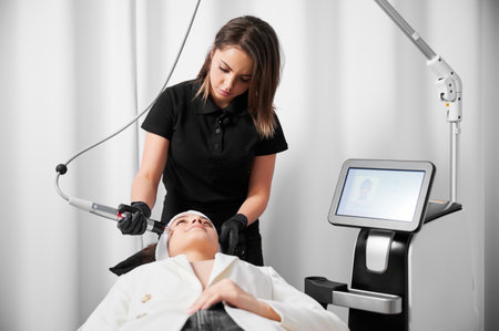Female cosmetologist using radiofrequency microneedling device while performing face lifting procedure. Young woman lying near cosmetology equipment and receiving skincare treatment.の写真素材