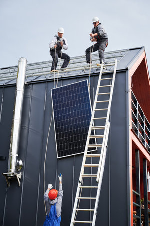 Men workers installing solar panel system on roof of house. Electricians in helmets lifting up photovoltaic solar module with help of ropes outdoors. Concept of alternative and renewable energy.の写真素材