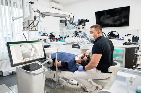 Dentist making X-ray, examining, inspecting problem, disease, holding equipment. Male doctor sitting on chair, fixing teeth, looking at screen. Concept of stomatology.の写真素材