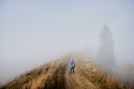 Photographer taking picture with professional camera while standing on grassy hill road with dense fog on backgroundの写真素材
