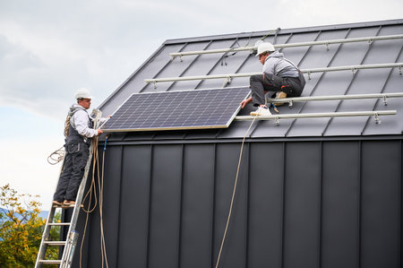 Mounters building photovoltaic solar module station on roof of house. Men electricians in helmets installing solar panel system outdoors. Concept of alternative and renewable energy.の写真素材