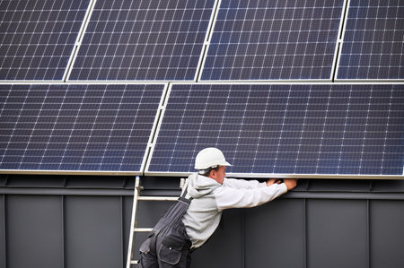 Electrician connecting cables while installing photovoltaic solar panels on roof of house. Back view of roofer in white helmet mounting of PV solar module on rooftop. Concept of renewable energy.の写真素材