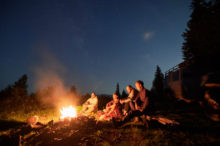 Night sky over mountain hill with car and male hikers near campfire. Group of travelers sitting near bonfire under beautiful blue sky with stars. Concept of night camping, hiking and travelling.の写真素材