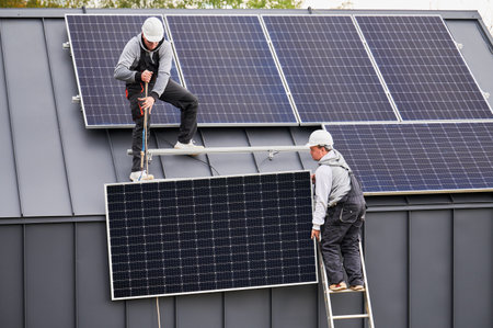 Men technicians installing solar panel system on roof of house. Workers in helmets lifting up photovoltaic solar module with help of ropes outdoors. Concept of alternative and renewable energy.の写真素材