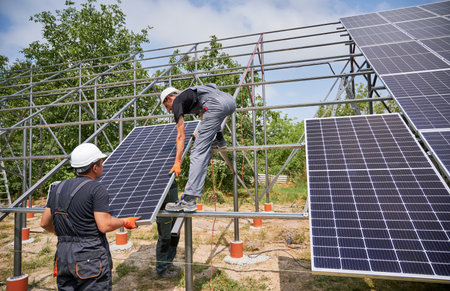 Workers installing solar panel on metal beams in field at sunny daytime. Renewable and ecological energy. Idea of environment safe. Modern technology and innovation. Men wearing workwear and helmetsの写真素材