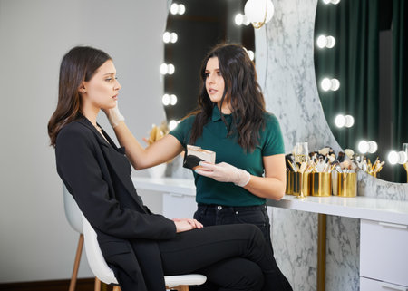 Stylish woman sitting on stool at dressing table while beauty specialist doing professional makeup in modern salon. Female beauty specialist applying cosmetics on woman face in visage studio.の写真素材