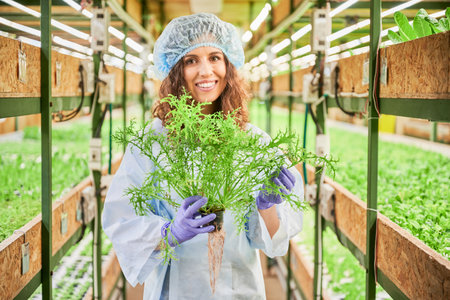Cheerful female gardener in disposable cap and garden gloves looking at camera and smiling while holding pot with green plant. Young woman with leafy plant in hands standing in greenhouse.の写真素材