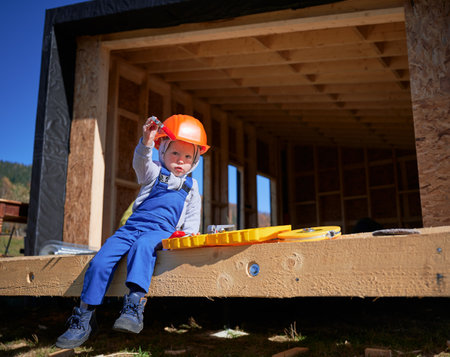 Boy toddler playing as builder on construction site. Child carpenter in orange helmet and blue overalls learning to build wooden frame house outdoor on sunny day. Carpentry and workshop concept.の写真素材