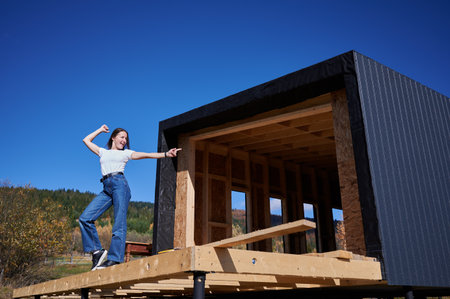 Happy woman on construction site standing on terrace at unfinished wooden frame house on sunny day, pointing at future building.の写真素材